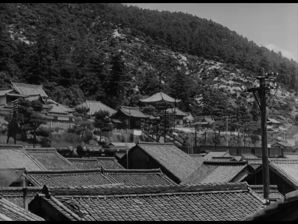一列冒烟的列车横向穿过层叠瓦屋顶后的轨道，后景可见林木与寺院。