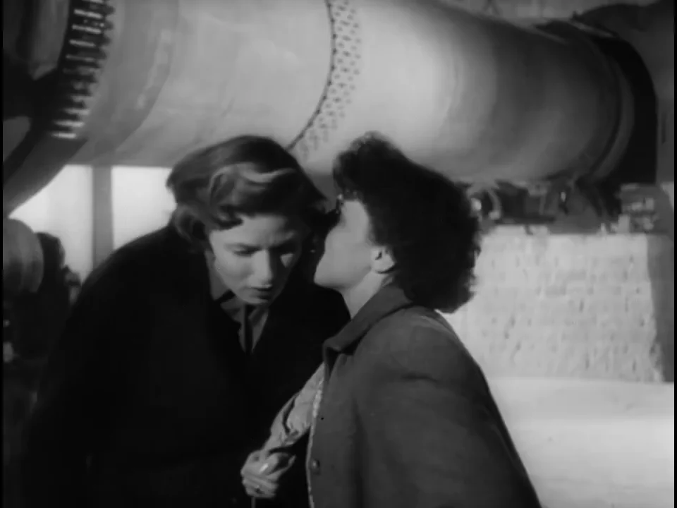 Irene leans toward a woman worker beneath a huge cylindrical machine in a passage crowded by pipes and machinery
