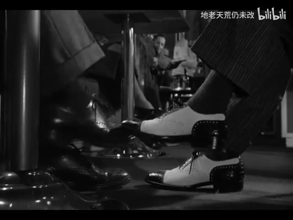火车包厢座位下，Guy 从左侧移来的深色鞋碰到 Bruno 右侧的双色便鞋；Bruno 另一只双色鞋停在右下方，画面没有显示二人的脸。