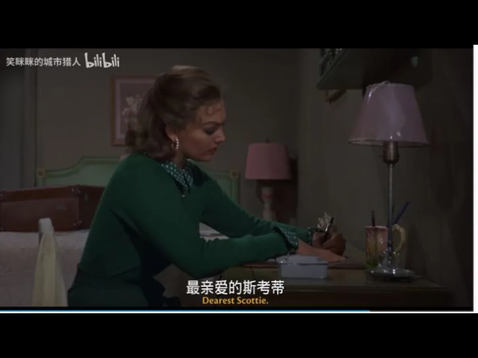 Judy 身穿绿色上衣坐在旅馆房间书桌前写信；画面底部的英文硬字幕可读为“Dearest Scottie”。